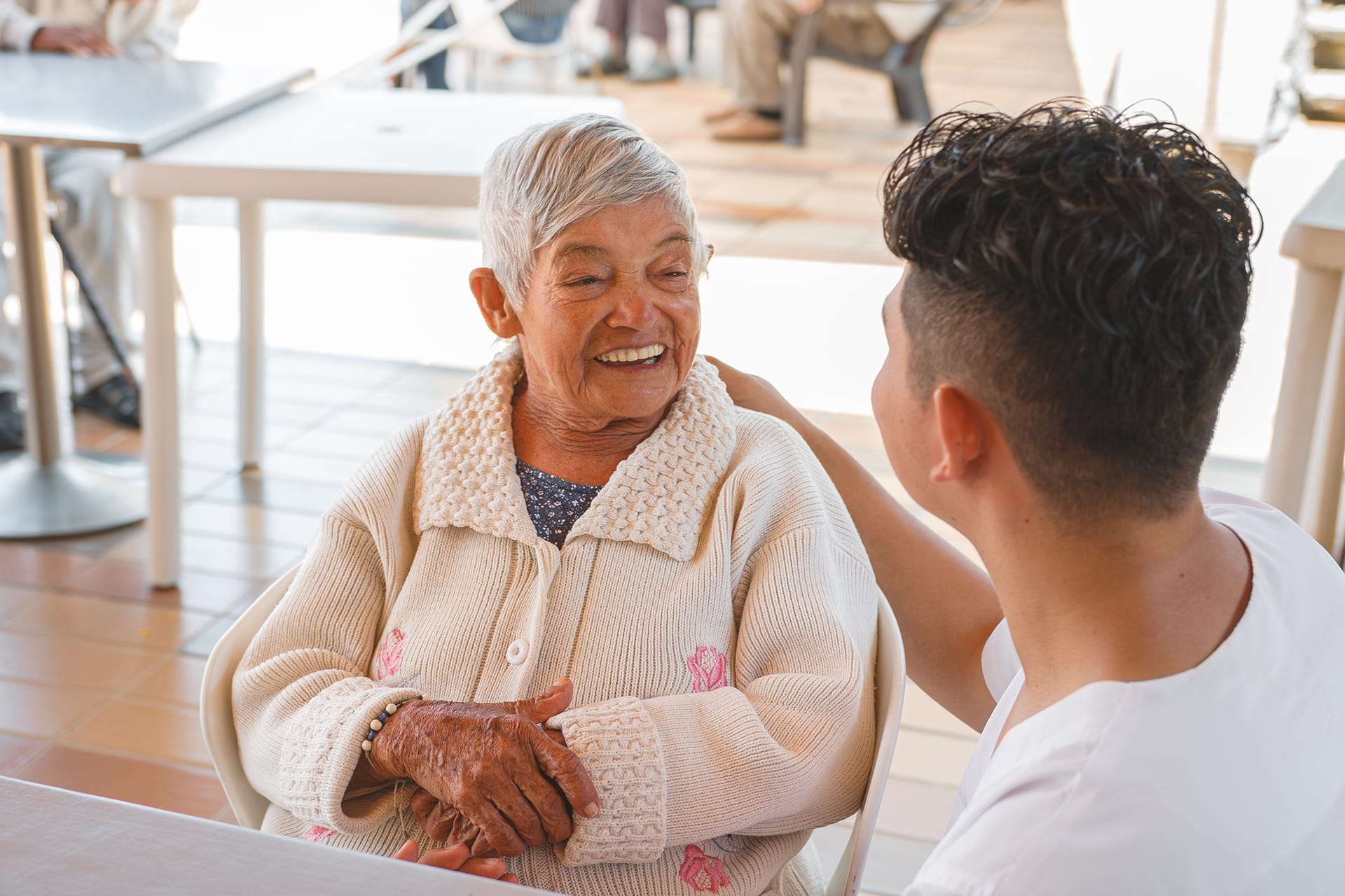 An elderly woman smiles warmly while talking with a caregiver seated across from her.