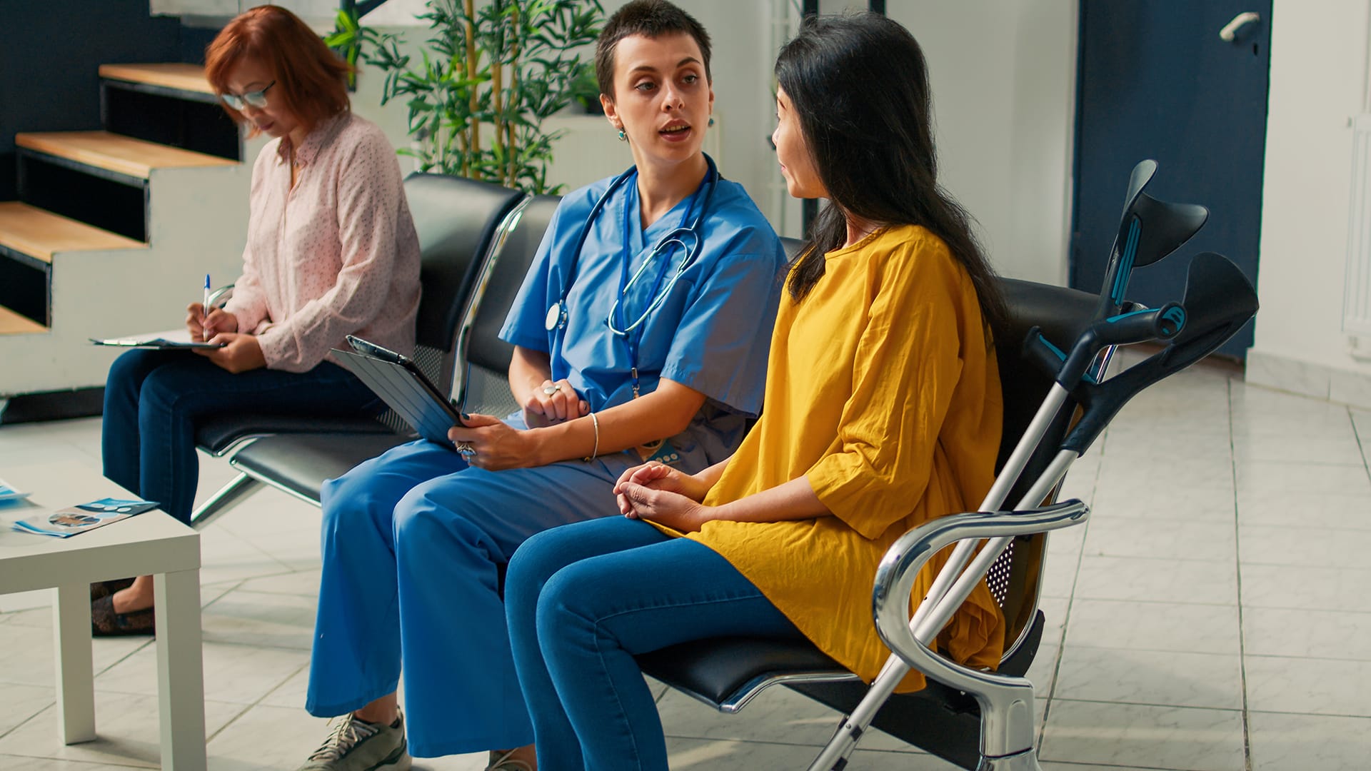 A nurse in scrubs talks with a woman on crutches in a waiting room, while another patient fills out paperwork nearby. The scene conveys a supportive healthcare environment.
